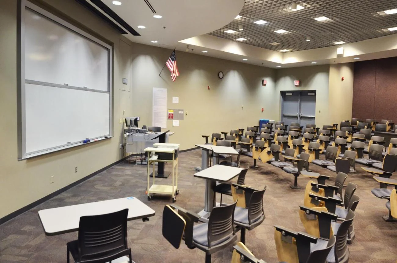 View of a classroom showing desks in rows a white board and a computer.